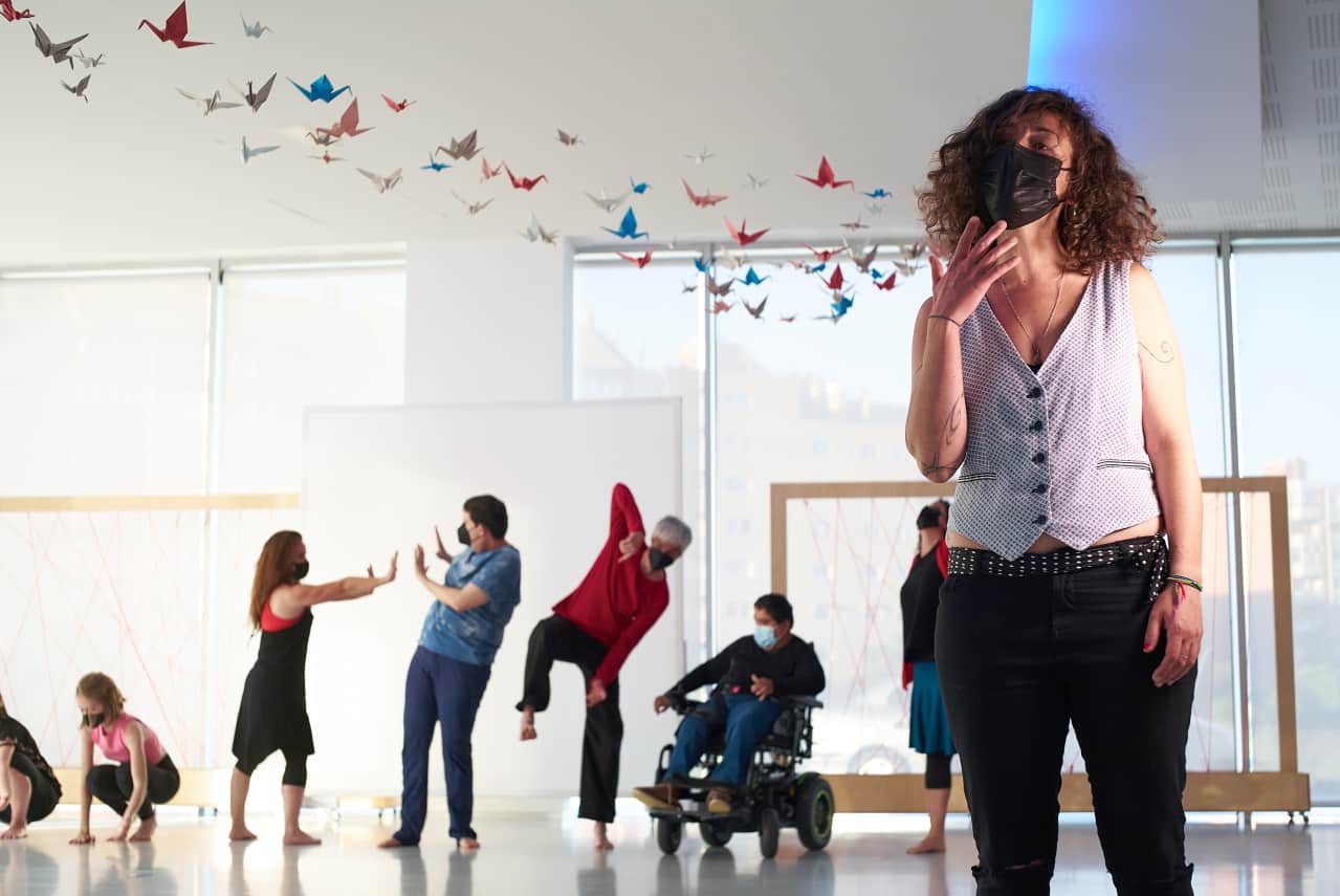 Mujer en primer plano actuando con varias personas de fondo bailando y colgadas del techo muchas gruyas de papel de muchos colores